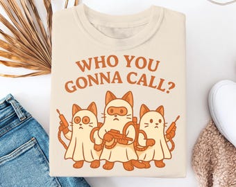 May include: A beige t-shirt with a graphic of three cartoon cats dressed as ghostbusters. The text "Who you gonna call?" is printed above the cats.