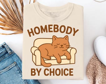 Peut inclure: Un t-shirt beige avec le texte "Homebody by Choice" en lettres brunes. Un chat orange dessin animé dort sur un canapé jaune.
