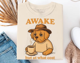Op de afbeelding: Een beige T-shirt met een cartoonillustratie van een hond die een hoed draagt en een kopje koffie vasthoudt. De tekst "AWAKE but at what cost" is op het T-shirt gedrukt.