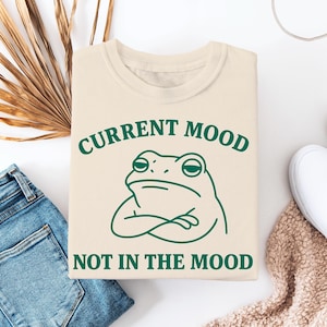 Op de afbeelding: Een beige sweatshirt met een groene kikker afbeelding en de tekst "CURRENT MOOD NOT IN THE MOOD".