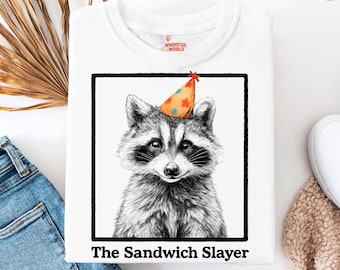 May include: White t-shirt featuring a raccoon wearing a party hat, framed in black. The text "The Sandwich Slayer" is printed below the raccoon illustration. The shirt has a crew neck and the brand "Whoopsie World" is visible.