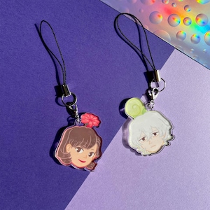 May include: Two acrylic charms with cartoon faces, each with a black lanyard. One charm features a brown-haired character with a pink flower, the other a white-haired character with a green swirl. The background is purple.