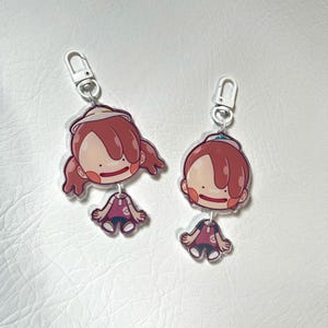 May include: Two acrylic keychains featuring a cartoon character with red hair, a white hat, and a pink and purple outfit. Each keychain has a silver clasp for attaching to keys or bags. The character's design is simple and cute.