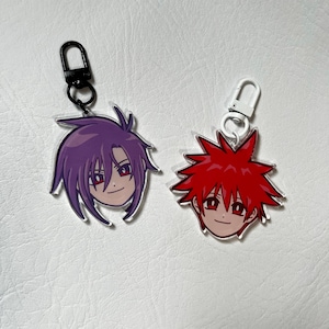 May include: Two anime-style character head keychains. One keychain features a purple-haired character with red eyes, the other a red-haired character with red eyes. Both have a smiling expression and are attached to a metal clasp.