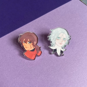 May include: Two acrylic pins featuring anime-style character portraits. One pin depicts a character with brown hair and a red outfit, the other with white hair and a black outfit. Both pins have a glossy finish and are set against a purple background.