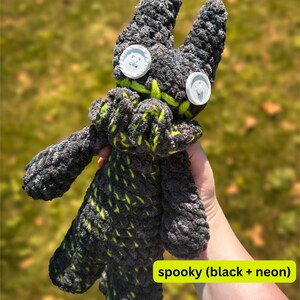 K&ouml;nnte beinhalten: Ein handgefertigtes Pl&uuml;schtier in Grau und Schwarz, akzentuiert mit neongr&uuml;nen N&auml;hten. Das Spielzeug hat gro&szlig;e Knopfaugen und einen gestickten Mund. Der Text "spooky (black + neon)" ist sichtbar. Das Pl&uuml;schtier wird vor einem verschwommenen gr&uuml;nen Hintergrund gehalten.