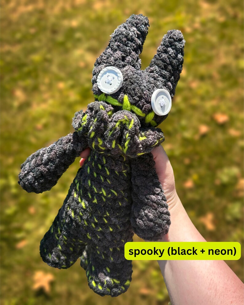 K&ouml;nnte beinhalten: Ein handgefertigtes Pl&uuml;schtier in Grau und Schwarz, akzentuiert mit neongr&uuml;nen N&auml;hten. Das Spielzeug hat gro&szlig;e Knopfaugen und einen gestickten Mund. Der Text "spooky (black + neon)" ist sichtbar. Das Pl&uuml;schtier wird vor einem verschwommenen gr&uuml;nen Hintergrund gehalten.