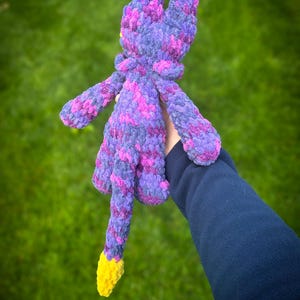 K&ouml;nnte beinhalten: Ein handgefertigtes Pl&uuml;schtier in Lila-, Rosa- und Gelbt&ouml;nen. Das Pl&uuml;schtier hat lange Ohren, ausgestreckte Arme und einen Schwanz mit gelber Spitze. Das Spielzeug besteht aus einem weichen, strukturierten Material und wird vor einem gr&uuml;nen Hintergrund gehalten.