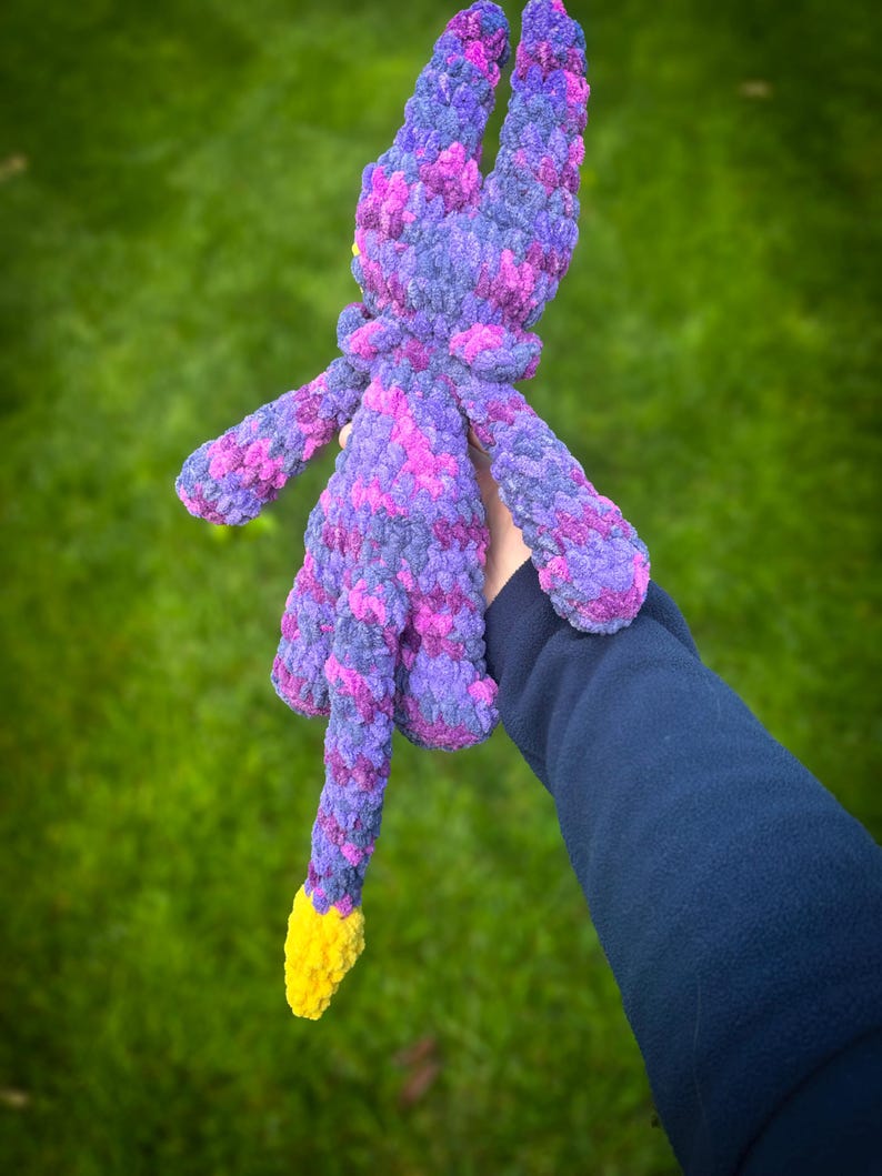 K&ouml;nnte beinhalten: Ein handgefertigtes Pl&uuml;schtier in Lila-, Rosa- und Gelbt&ouml;nen. Das Pl&uuml;schtier hat lange Ohren, ausgestreckte Arme und einen Schwanz mit gelber Spitze. Das Spielzeug besteht aus einem weichen, strukturierten Material und wird vor einem gr&uuml;nen Hintergrund gehalten.