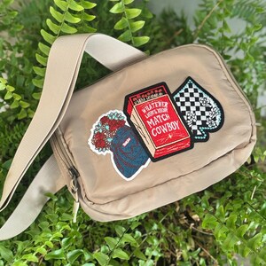 May include: Tan crossbody bag with a beige strap. The bag features three embroidered patches: a bouquet, a matchbox with the text "WHATEVER LIGHTS YOUR MATCH COWBOY", and a black and white checkered spade with floral accents.
