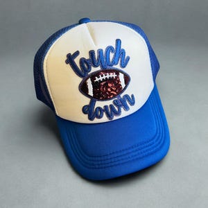 May include: A blue and white trucker hat with the words "Touch Down" in script. The hat features a football design with sequin embellishments. The brim and front panel are blue, while the crown is white.