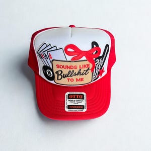 May include: Red and white trucker hat with the text "SOUNDS LIKE BULLSHIT TO ME." The hat features a red brim and mesh back, with a white front panel displaying playing cards, a pool ball, and a red bow.