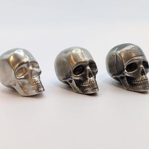 May include: Three small, silver-colored skull figurines are arranged in a row against a white background. The skulls have detailed facial features, including eye sockets, a nose cavity, and teeth. The skulls appear to be made of metal.