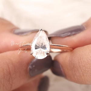 Peut inclure: Une bague de fiançailles en diamant de forme poire sertie sur une bande en or rose. La bague est sur un doigt avec un vernis à ongles gris.