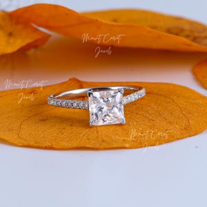 May include: A silver engagement ring with a square-shaped diamond and a band of small diamonds. The ring is on a bed of orange leaves.