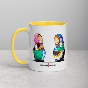 Pode incluir: Caneca de cerâmica branca com borda amarela. A caneca apresenta uma ilustração colorida de duas bonecas russas de encaixe, ou bonecas Matryoshka, com o texto "Matryoshka Emotions" na parte inferior.