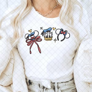 May include: White t-shirt featuring three cartoon character headbands: one with a large pink bow, one with Donald Duck's face, and one with a smaller red bow. The design includes stars and hearts.