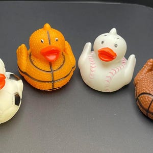 Op de afbeelding: Vier rubberen badeenden, elk ontworpen om op een andere sport te lijken. De eenden zijn wit, oranje en bruin, met details zoals een voetbal, basketbal en honkbal. Ze hebben oranje snavels.