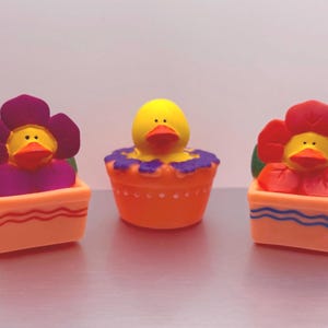 Puede incluir: Tres figuras decorativas coloridas. Dos tienen forma de flor con caras de pato, y una es un pato en una maceta. Las figuras en forma de flor tienen pétalos rojos y morados, y el pato tiene un cuerpo amarillo y un pico naranja.
