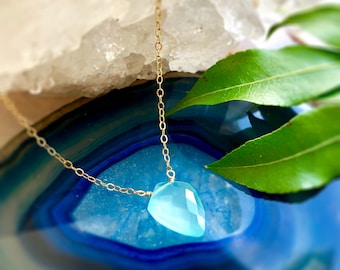 May include: A delicate gold chain necklace featuring a faceted, light blue gemstone pendant. The pendant is teardrop-shaped and hangs from a fine gold chain. The necklace is displayed on a blue and white surface with green leaves.