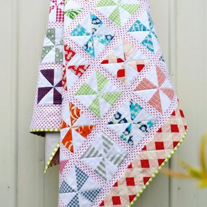 May include: A white quilt with a patchwork design featuring colourful pinwheels. The quilt has a red and white gingham border and a green trim.