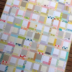 May include: A patchwork quilt with a variety of colorful fabrics and patterns. The quilt is made up of squares and rectangles, and the squares are outlined in white. The quilt is a light beige color with pops of pink, blue, green, yellow, and orange.