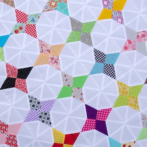 May include: A close-up shot of a handmade quilt with a repeating pattern of white geometric shapes. The quilt features colourful star-shaped patches in various patterns and colours, including red, yellow, green, and blue, set against a white background.