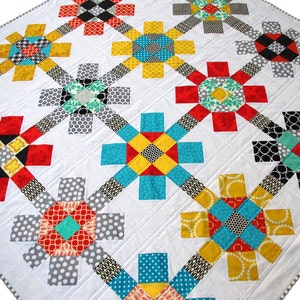 Op de afbeelding: Een wit quilt met een herhalend patroon van kleurrijke, geometrische bloemenvormen. Het quilt is gemaakt van verschillende stoffen, waaronder rood, geel, blauw, zwart en wit.