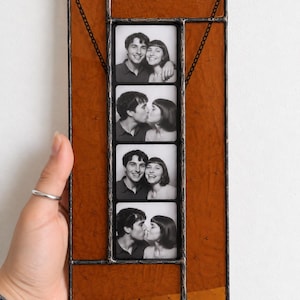 May include: A rectangular stained glass frame with a dark metal border and amber-colored glass panels. The frame holds a vertical strip of four black and white photo booth pictures showing two people smiling and kissing in a sequence of poses.