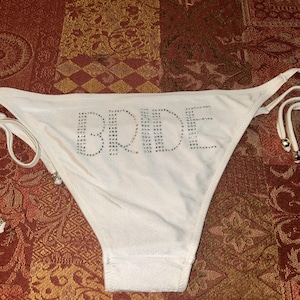 May include: A white bikini bottom featuring the word BRIDE spelled out in small, clear rhinestones across the front. The garment has adjustable side ties with thin strings and small silver-toned metal beads at the ends of each tie.