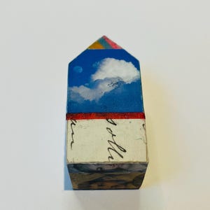 Peut inclure: Petite œuvre d'art en forme de maison avec un ciel bleu et des nuages. Le sommet est peint avec un motif arc-en-ciel. La partie inférieure comporte du texte manuscrit et une bande rouge. La base est un collage d'images.
