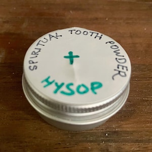 Könnte beinhalten: Kleine, silberfarbene Dose mit weißem Deckel. Auf dem Deckel steht in einem Kreis "SPIRITUAL TOOTH POWDER", ein grünes Kreuz und das Wort "HYSSOP" in Grün. Die Dose steht auf einer Holzoberfläche.