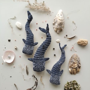 May include: Three navy blue whale shark plush toys with white polka dots, arranged with seashells, dried seaweed, and small stones on a light background. The toys have fins and curved tails.