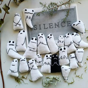 Op de afbeelding: Een verzameling kleine, spookachtige pluche figuren gemaakt van witte stof met zwart geborduurde gelaatstrekken en ledematen. Eén figuur is volledig zwart. Ze zijn gerangschikt rond een rechthoekige kaart met het woord SILENCE in grijs.