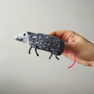 May include: A hand holds a small, gray and white embroidered opossum with a pink tail. The opossum is a handmade, needle-felted toy.