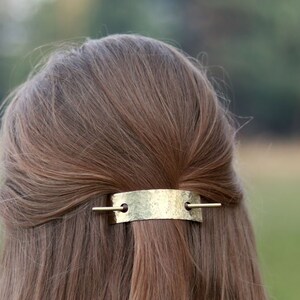 May include: A gold-toned metal hair clip with a hammered texture. The clip is in the shape of a rectangle with rounded edges and has two small holes on either side. The clip is being used to hold back a woman's long brown hair.