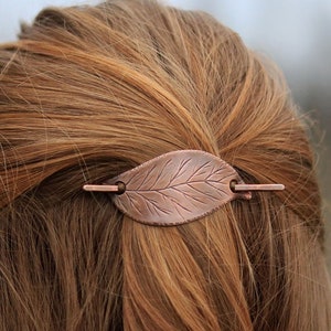 以下が含まれることがあります： 葉の形をした銅色のヘアバレッタで、詳細な葉脈があります。 バレッタは茶色の髪に固定されており、シンプルなピンのデザインです。 自然にインスパイアされたヘアアクセサリーです。