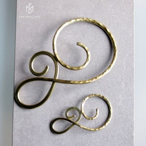 May include: Two gold metal hair clips in the shape of a swirl. The larger clip is on the left and the smaller clip is on the right. Both clips have a hammered texture.