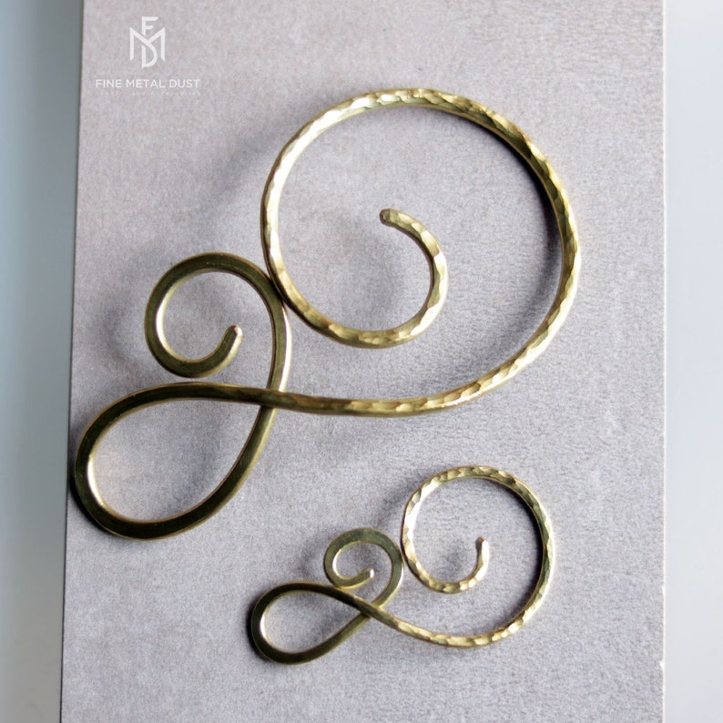 May include: Two gold metal hair clips in the shape of a swirl. The larger clip is on the left and the smaller clip is on the right. Both clips have a hammered texture.