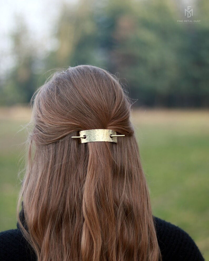 May include: A gold-toned metal hair clip with a hammered texture. The clip is in the shape of a rectangle with rounded edges and has two small holes on either side. The clip is being used to hold back a woman's long brown hair.