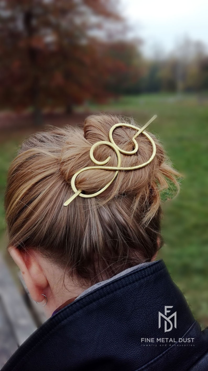 May include: Gold hair stick with a decorative swirl design. The stick is being used to hold a woman's hair in a bun. The hair stick is made by Fine Metal Dust Jewelry and Accessories.