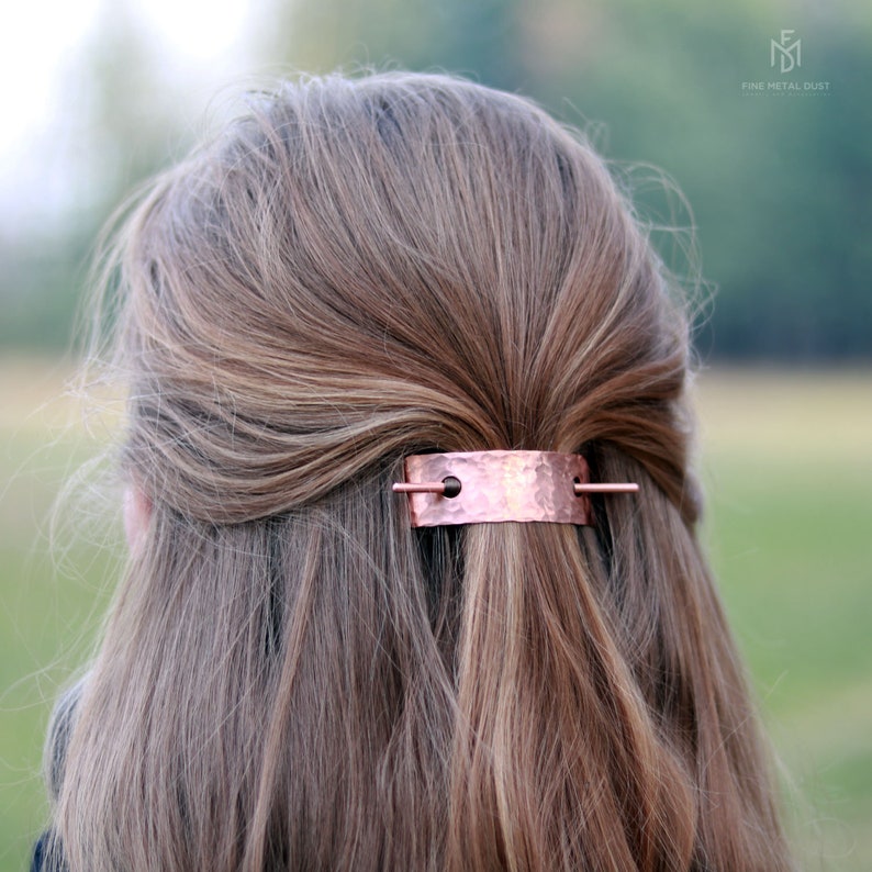 May include: A hammered copper hair clip with a thin metal bar that slides through two holes. The clip is worn in a woman's hair.