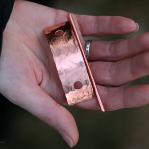 May include: A hammered copper hair stick with a smooth, rounded end. The stick is held in a hand, and the copper has a warm, reddish hue.