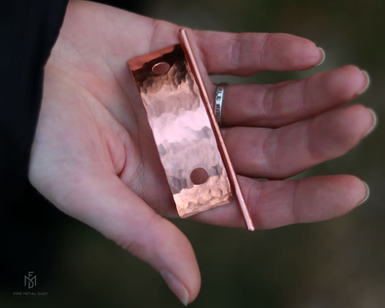 May include: A hammered copper hair stick with a smooth, rounded end. The stick is held in a hand, and the copper has a warm, reddish hue.