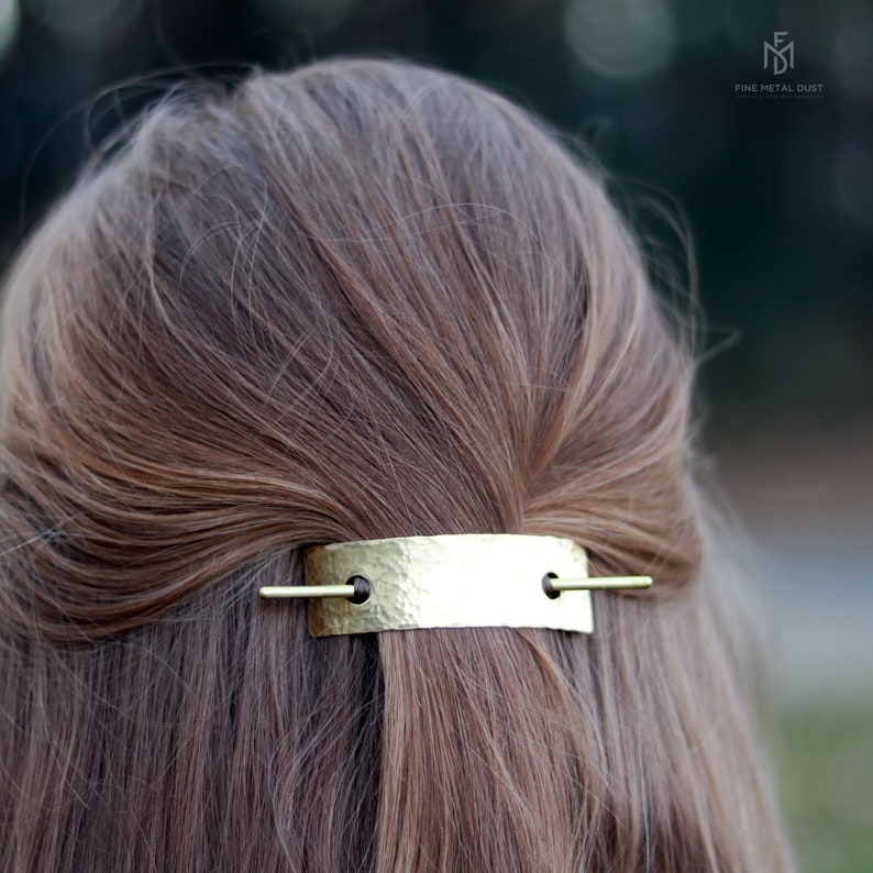 May include: A gold hammered metal bar hair clip with two small holes and a thin gold bar extending from each hole. The clip is in a person's hair.