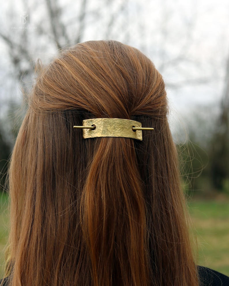May include: A gold-toned hair clip with a hammered texture and a straight bar. The clip is in a woman's brown hair.