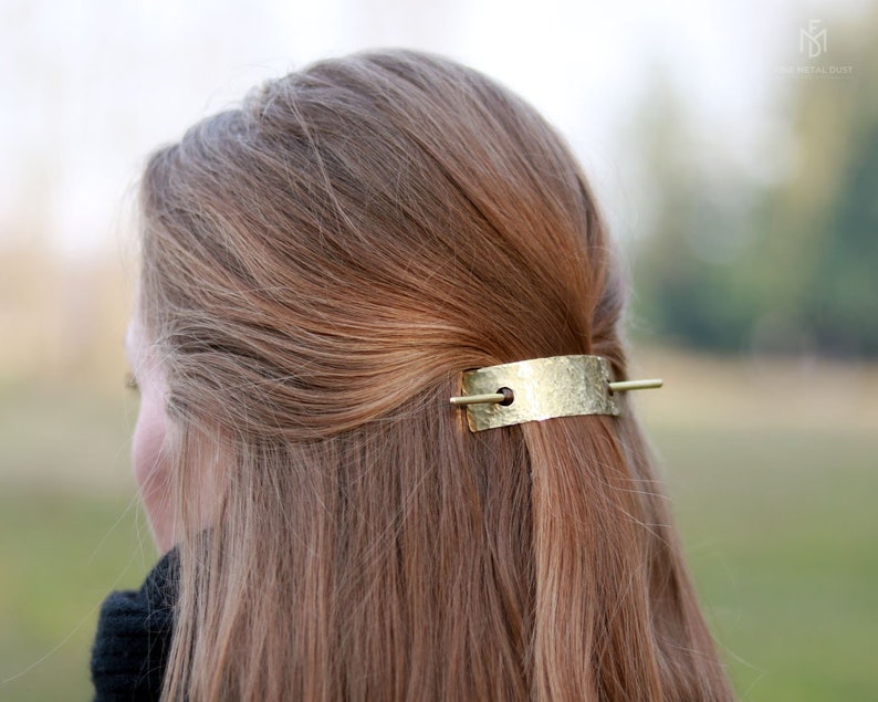 May include: A gold-toned hair clip with a hammered texture and a thin, straight bar. The clip is in a person's hair.