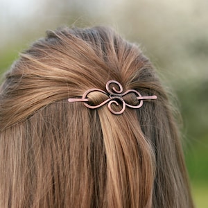 May include: Copper-colored hair barrette with an intricate, swirling design. The barrette is made of wire and is inserted into the hair. The hair is a light brown color and is styled in a half-up, half-down style. The barrette is the focal point of the image.