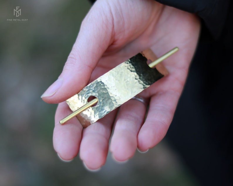 May include: A gold hammered metal hair stick with a smooth, round pin. The stick is rectangular and has a hammered texture.