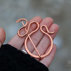 Pode incluir: Um broche em forma de cobra de cor cobre com um alfinete. O broche tem a forma de uma cobra com a cauda enroscada em torno da cabeça. O alfinete está preso ao corpo da cobra.
